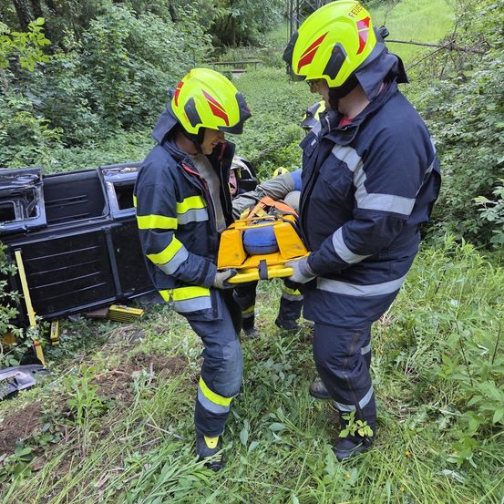 Zwei Feuerwehrleute befinden sich in einem Feld in der Nähe eines umgekippten Fahrzeugs. Sie halten ein gelbes Werkzeug und tragen Helme und Handschuhe.