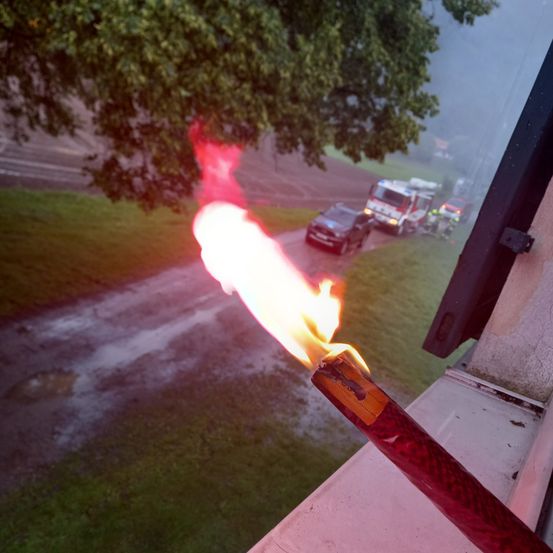 Eine Fackel mit einer Flamme brennt hell im Regen. Ein Feuerwehrauto und andere Fahrzeuge sind auf einer Straße im Hintergrund geparkt.
