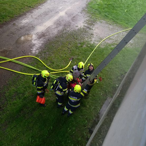 Feuerwehrleute in gelben Uniformen retten eine Person auf einem nassen Grasfeld mit Schläuchen drumherum.