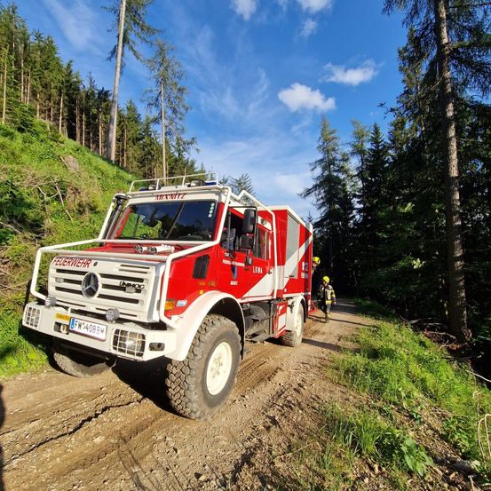 Ein rot-weißer Feuerwehrwagen fährt auf einer unbefestigten Straße, wobei zwei Feuerwehrleute an der Seite gehen. Die Straße ist von hohen Bäumen umgeben.