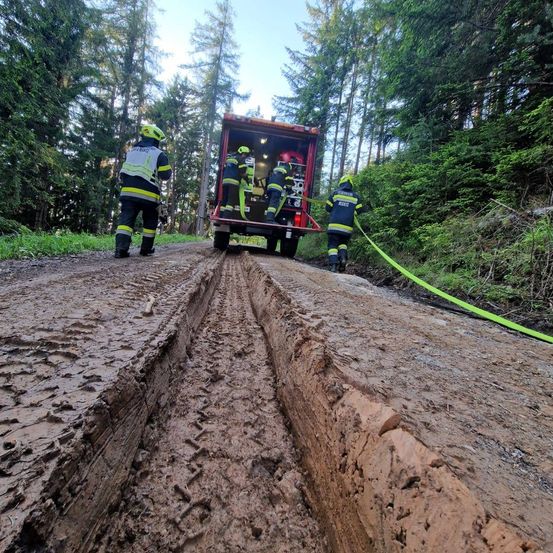 Feuerwehrleute in Uniform und Helmen sind auf einer schlammigen Straße, zwei von ihnen sind auf einem LKW, während die anderen beiden eine Schläuche ziehen.