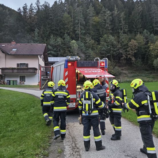 Feuerwehrleute in Schutzausrüstung versammeln sich um einen roten Feuerwehrwagen. Ein Haus mit Solarmodulen im Hintergrund.