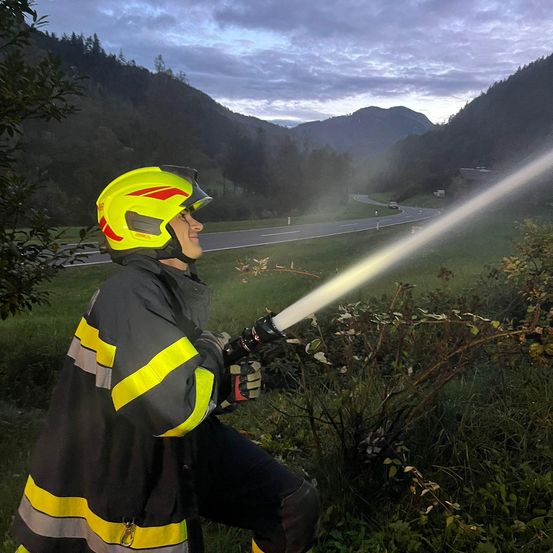 Ein Feuerwehrmann in einem gelben Helm und reflektierender Kleidung sprüht Wasser aus einem Schlauch, mit einer kurvenreichen Straße und Bergen im Hintergrund.