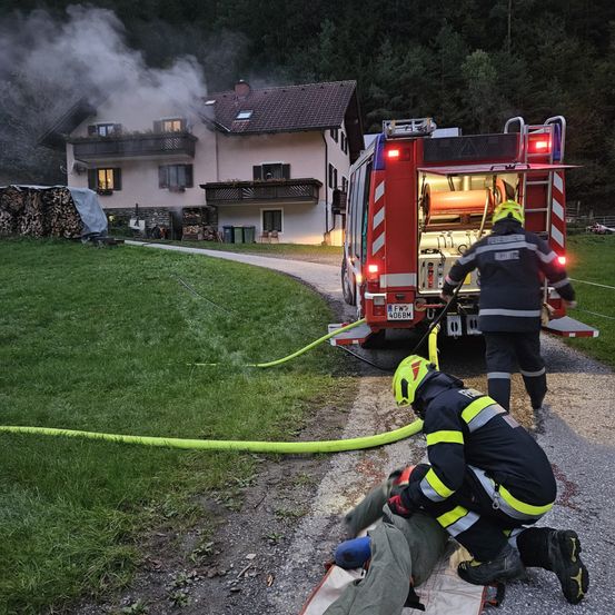 Feuerwehrleute kümmern sich um eine Person auf einer Trage, während ein Feuerwehrwagen mit einem Schlauch verbunden ist, wobei ein Haus Rauch abgibt.