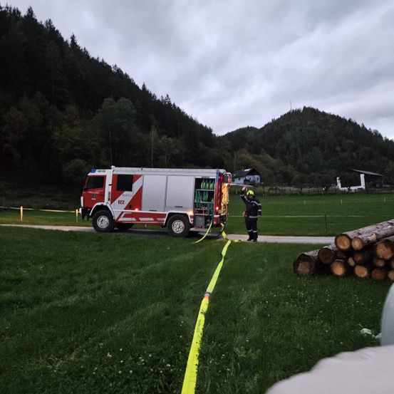 Ein Feuerwehrmann steht neben einem rot-weißen Feuerwehrwagen auf einem Rasenfeld mit einer gelben Warnlinie, mit Bergen und einem Haus im Hintergrund.