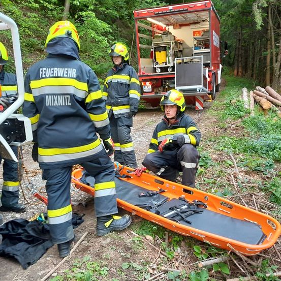 Feuerwehrleute in Schutzkleidung bereiten eine orangefarbene Trage in einem Waldgebiet vor. Ein roter Feuerwehrwagen ist in der Nähe geparkt, und Holzstämme sind auf dem Boden verstreut.