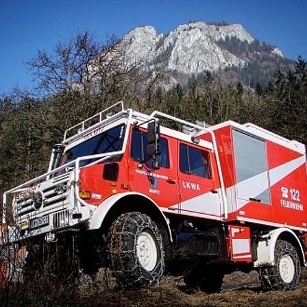 Ein rot-weißer Feuerwehrwagen mit Ketten an den Reifen steht in einem bewaldeten Gebiet mit einem Berg im Hintergrund.