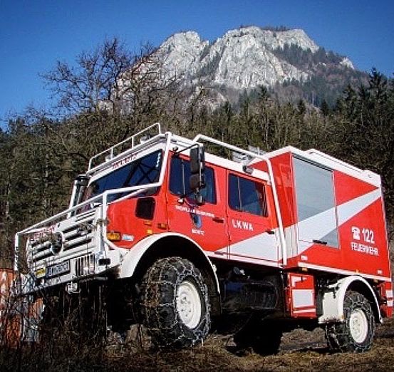 Ein rot-weißer Feuerwehrwagen mit Ketten an den Reifen steht in einem bewaldeten Gebiet mit einem Berg im Hintergrund.