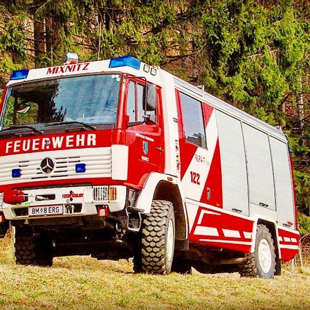 Ein rot-weißer Feuerwehrwagen mit der Aufschrift 'Feuerwehr' steht auf einer Wiese mit Bäumen im Hintergrund.