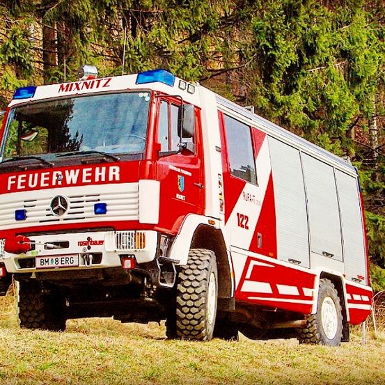Ein rot-weißer Feuerwehrwagen mit der Aufschrift 'Feuerwehr' steht auf einer Wiese mit Bäumen im Hintergrund.