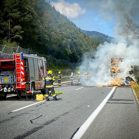 Feuerwehrleute bekämpfen ein Feuer auf einer Straße mit einem brennenden Fahrzeug, umgeben von Bäumen und Bergen.