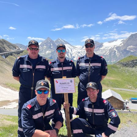 Fünf Männer in Uniformen posieren für ein Foto vor einer Berglandschaft. Sie stehen auf einem Rasen mit Bergen im Hintergrund.