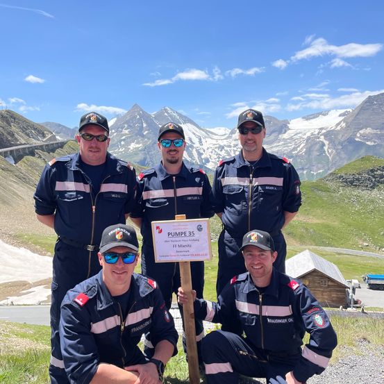 Fünf Männer in Uniformen posieren für ein Foto vor einer Berglandschaft. Sie stehen auf einem Rasen mit Bergen im Hintergrund.