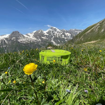 Ein grünes Übungsband mit der Aufschrift 'FF MIXWITZ' liegt auf einer Wiese mit Blumen und verschneiten Bergen im Hintergrund.