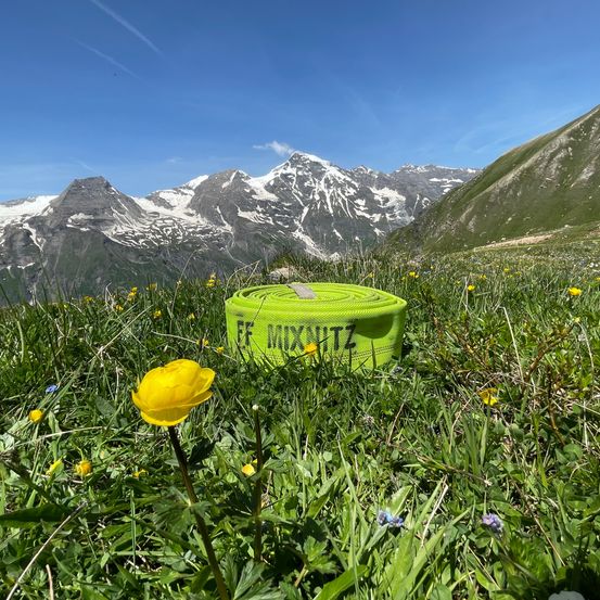 Ein grünes Übungsband mit der Aufschrift 'FF MIXWITZ' liegt auf einer Wiese mit Blumen und verschneiten Bergen im Hintergrund.