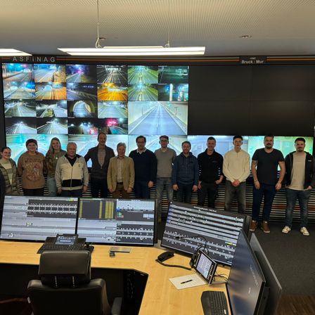 Eine Gruppe von Menschen steht vor einem großen Monitor, der mehrere Verkehrskameras anzeigt. Sie befinden sich in einem Büro mit Computerbildschirmen, Stühlen und einem Schreibtisch. An der Wand befindet sich ein großer Bildschirm mit den Texten 'ASFINAIG' und 'Bruck/Mur'.