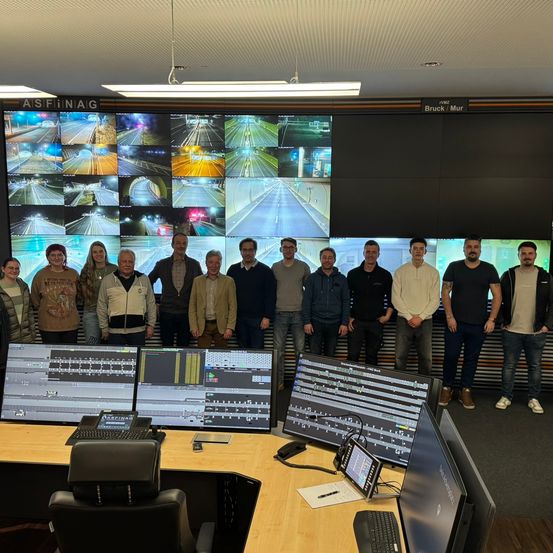 Eine Gruppe von Menschen steht vor einem großen Monitor, der mehrere Verkehrskameras anzeigt. Sie befinden sich in einem Büro mit Computerbildschirmen, Stühlen und einem Schreibtisch. An der Wand befindet sich ein großer Bildschirm mit den Texten 'ASFINAIG' und 'Bruck/Mur'.