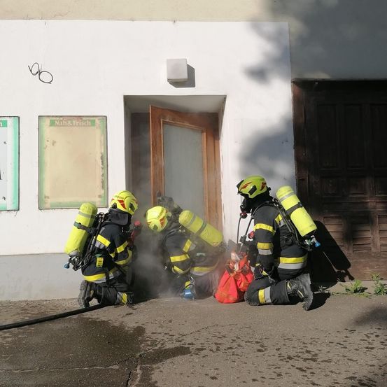 Drei Feuerwehrleute in gelben Helmen und gelben Uniformen knien auf dem Bürgersteig. Sie scheinen sich auf einen Notfalleinsatz vorzubereiten.