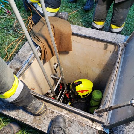 Ein Feuerwehrmann in einem gelben Helm ist in einem Brunnen und trägt Schutzausrüstung. Ein Seil und eine Metallleiter befinden sich neben dem Brunnen. Andere Feuerwehrleute stehen in der Nähe.
