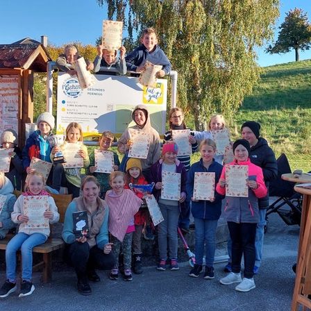 Kinder und Erwachsene posieren für ein Foto mit Urkunden vor einem Schild und einem Gebäude unter blauem Himmel.