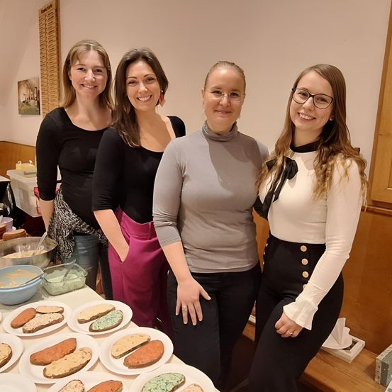 Vier Frauen stehen gemeinsam an einem Tisch mit Tellern voller Sandwiches. Sie lächeln und scheinen für ein Foto zu posieren. Der Tisch ist mit verschiedenen Sandwiches gefüllt. Hinter ihnen befindet sich eine Wand mit einem Bilderrahmen und einer weißen Tischdecke.