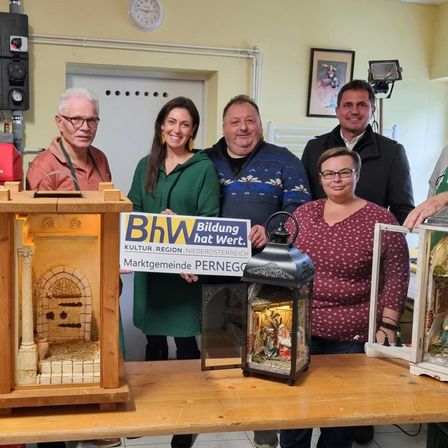 Fünf Personen stehen in einem Raum um einen Tisch mit beleuchteten Displays. Sie lächeln und posieren für ein Foto. Ein Schild mit dem Text BHV Bildung hat Wert. ist prominent angebracht.