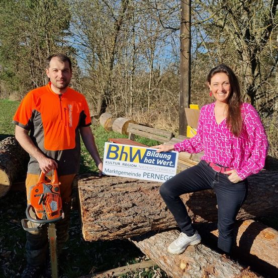 Zwei Personen stehen auf einem Holzhaufen. Ein Mann hält eine Kettensäge und eine Frau hält ein Schild mit der Aufschrift 'BhW'. Sie lächeln.