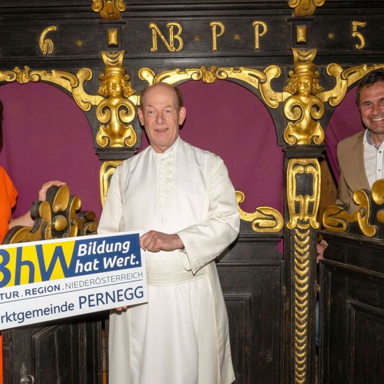 Ein Priester in einer weißen Robe hält ein blau-gelbes Schild mit den Worten 'Bildung hat Wert'. Zwei Männer, einer in einem braunen Mantel, stehen hinter ihm. Sie stehen vor einer dekorativen Wand mit einem Holzrahmen und goldenen Akzenten.