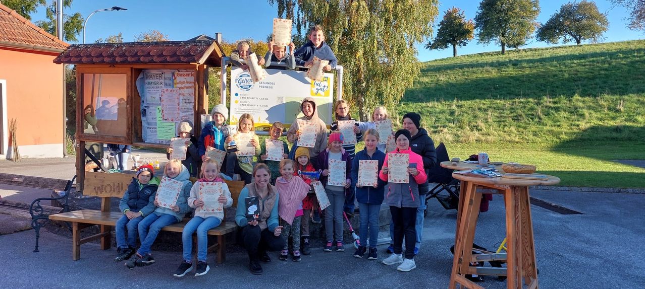 Eine Gruppe von Kindern und Erwachsenen posiert für ein Foto mit Urkunden und Auszeichnungen in der Hand. Sie stehen und sitzen auf einer Bank draußen, mit einem Banner dahinter.