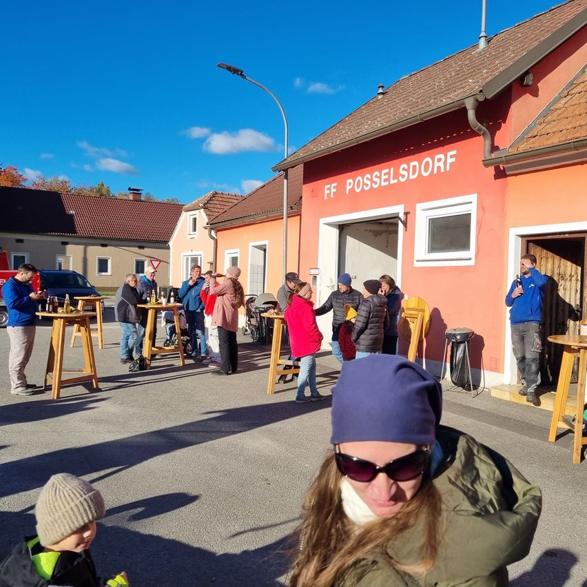 Eine Gruppe von Menschen steht vor dem Gebäude der FF Posselsdorf, einige sitzen an Tischen, andere gehen, und ein Mülleimer steht am Eingang.