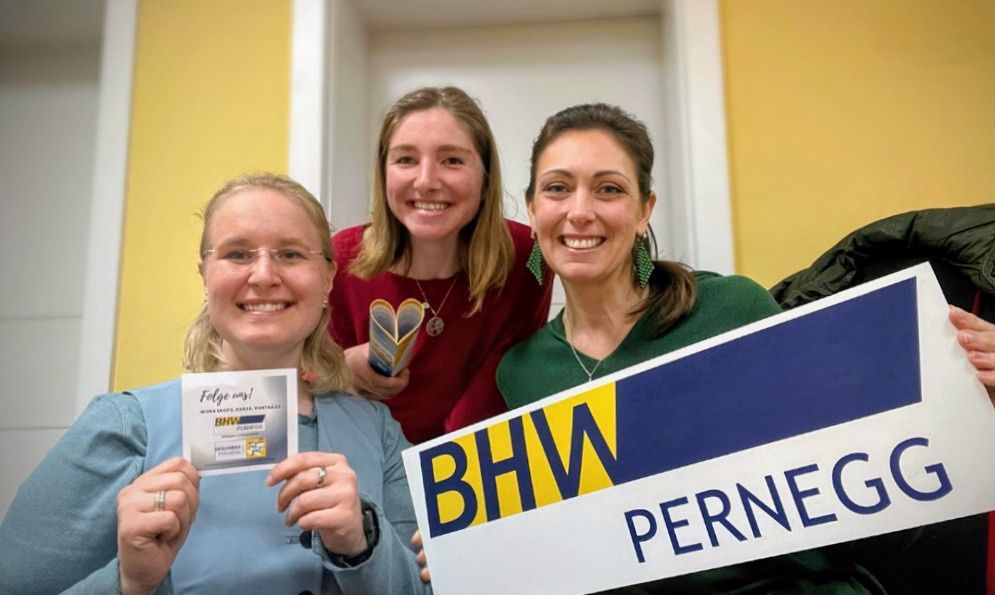 Drei Frauen lächeln für die Kamera, eine hält ein Schild mit 'BHW PERNEG', eine andere eine Karte mit einem herzförmigen Logo und die dritte ein Buch.