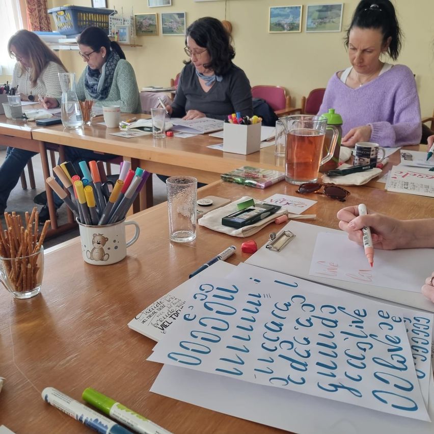 Eine Gruppe von Frauen sitzt an einem Tisch und nimmt an einem Kalligrafie-Workshop teil. Der Tisch ist mit verschiedenen Stiften, Markern und Papier gefüllt. Jede Frau konzentriert sich auf ihre eigene Kalligrafie-Übung.