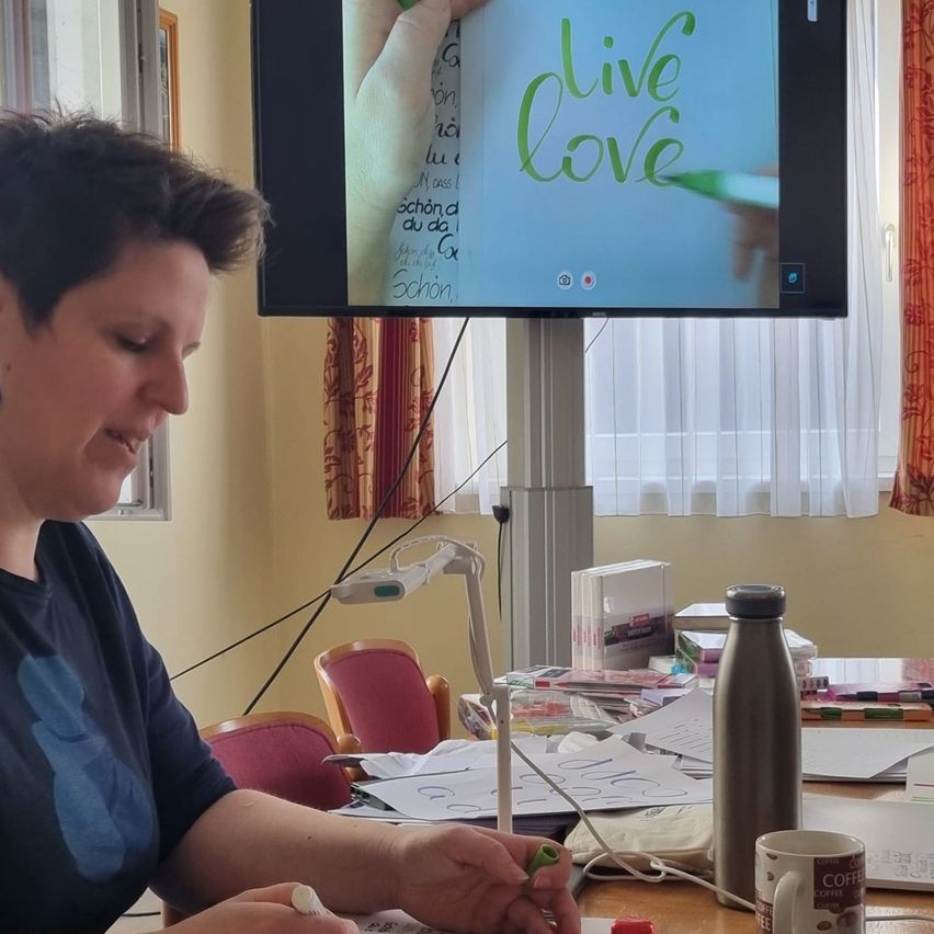 Eine Frau sitzt und zeichnet auf eine Tafel mit der Aufschrift 'live love', die auf einem Monitor hinter ihr angezeigt wird. In der Nähe befindet sich ein Schreibtisch mit einer Kaffeetasse, einer Flasche und verschiedenen Gegenständen.