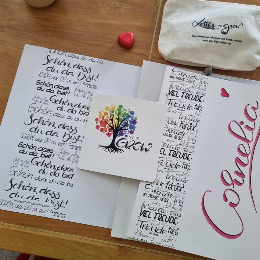 Handgeschriebene Kalligraphiekarten, ein Baumlogo und ein kleines Herz liegen auf einem Holztisch. Ein Beutel mit dem Text 'Buchstabe und wachsen' befindet sich neben den Karten.