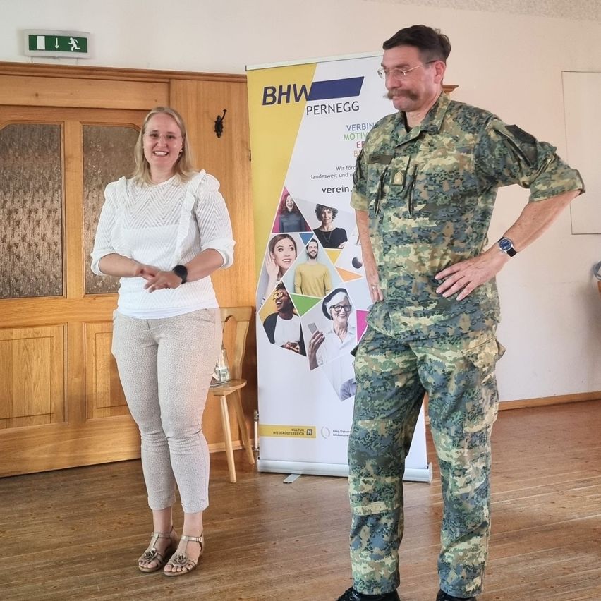 A man in a camouflage uniform stands next to a smiling woman in a white blouse in a room with wooden floors and a wooden door. Behind them is a banner with multiple faces and text.