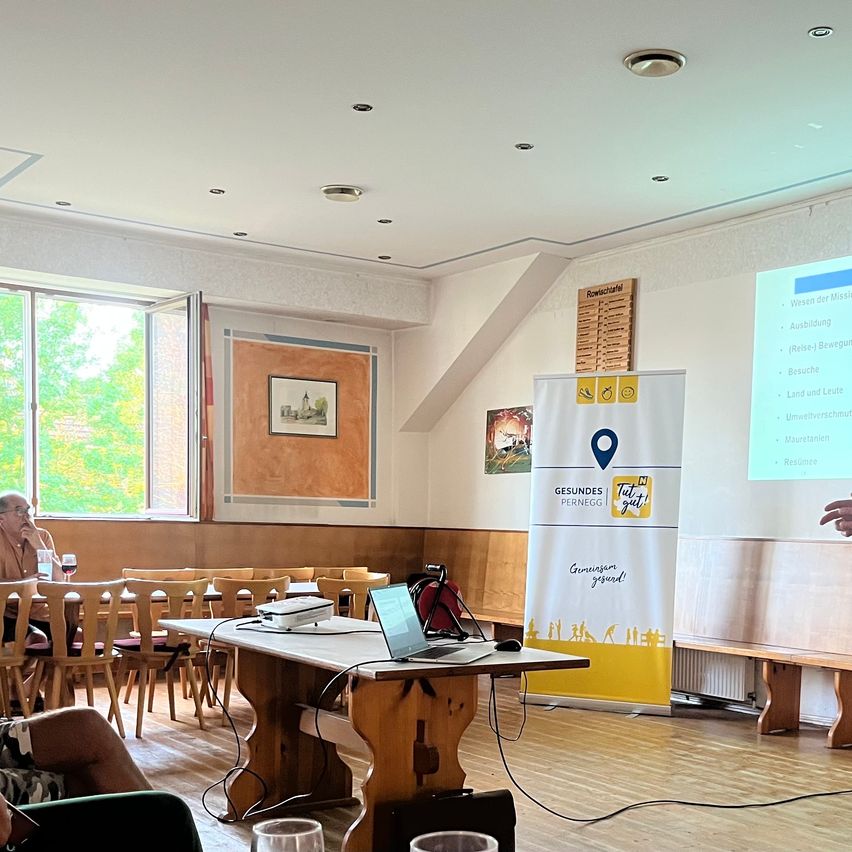 A man giving a presentation in a room with a projector screen, a laptop on a table, and a banner on the wall.