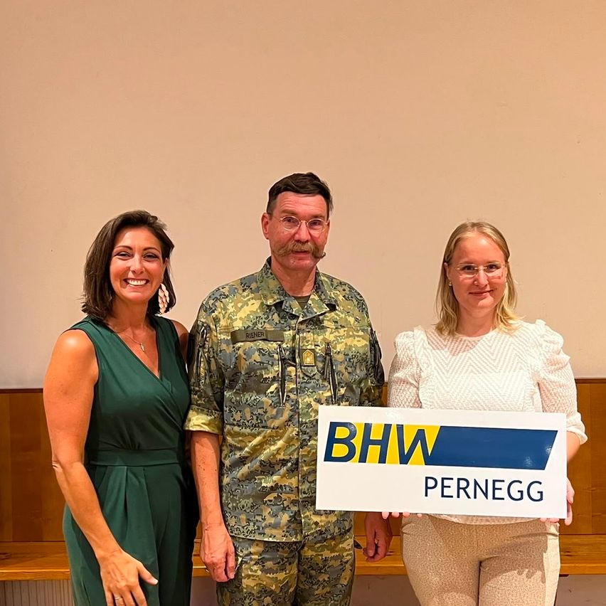 Three people stand together, smiling. A man in a military uniform holds a sign that says 'BHW Pernegg'. A woman on the left wears a green dress, and another woman on the right wears a white blouse.