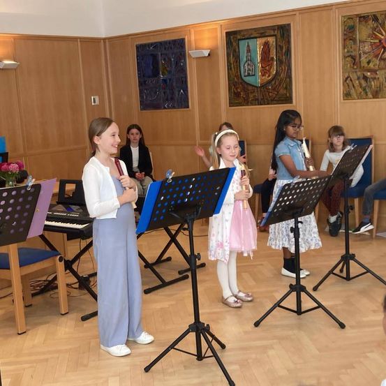 Mehrere junge Mädchen stehen in einem Raum, halten Musikinstrumente und lächeln. Dahinter sitzen einige, während andere stehen. Musikständer stehen vor ihnen und ein Klavier dahinter. An der Wand hängen Gemälde.