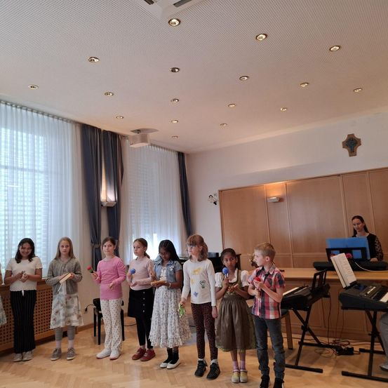 Eine Gruppe von Kindern in einem Raum mit Musikinstrumenten, einem Klavier und einer Frau. Die Kinder halten bunte Maracas und scheinen sich auf eine Aufführung vorzubereiten.
