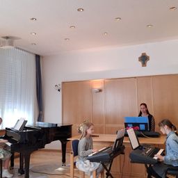 Drei Kinder spielen Klaviere in einem Raum mit Holzwänden und einer weißen Decke mit Kreuzsymbol. Eines spielt ein Flügel, die anderen Keyboards. Eine Frau steht hinter einem Schreibtisch mit Laptop und Blumen.