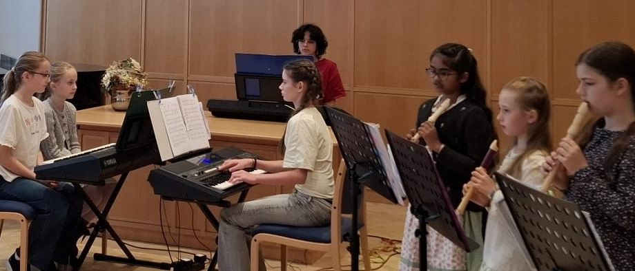 Eine Gruppe junger Mädchen übt gemeinsam Musik. Zwei spielen Keyboard, zwei spielen Flöten und eine spielt Klavier. Eine Frau steht hinter ihnen.