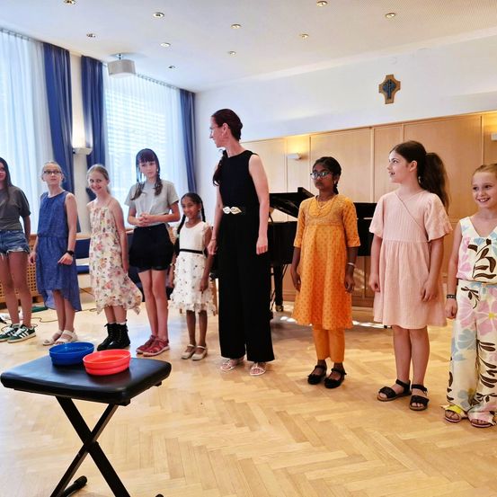 Eine Gruppe von Mädchen und eine Frau in Schwarz stehen in einem Raum und schauen sich gegenseitig an. Ein Klavier ist im Hintergrund.