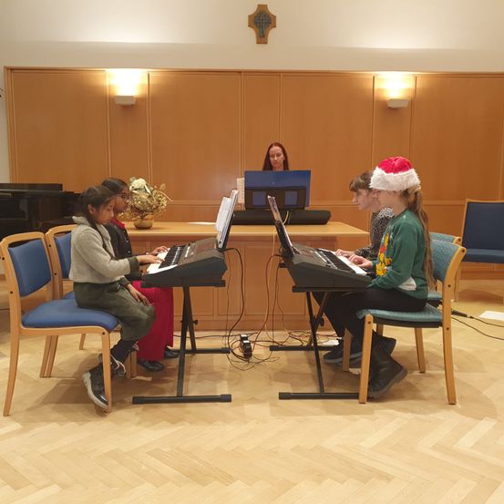 Fünf Kinder spielen elektrische Keyboards in einem Raum. Ein Mädchen trägt einen Weihnachtsmannhut. Hinter ihnen sitzt eine Frau an einem Schreibtisch mit Laptop.