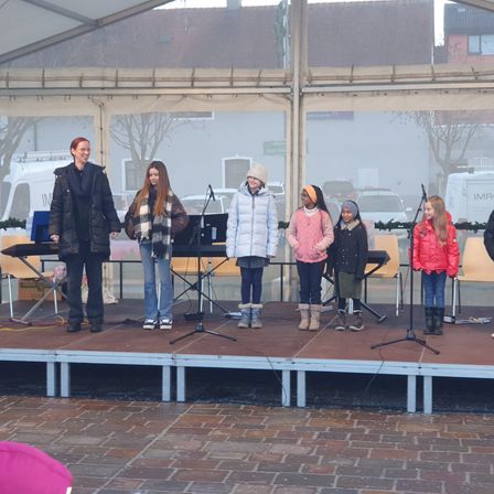 Eine Gruppe von Kindern steht auf einer Bühne unter einem Zelt mit einer erwachsenen Frau. Sie haben Musikinstrumente und scheinen sich auf eine Aufführung vorzubereiten.