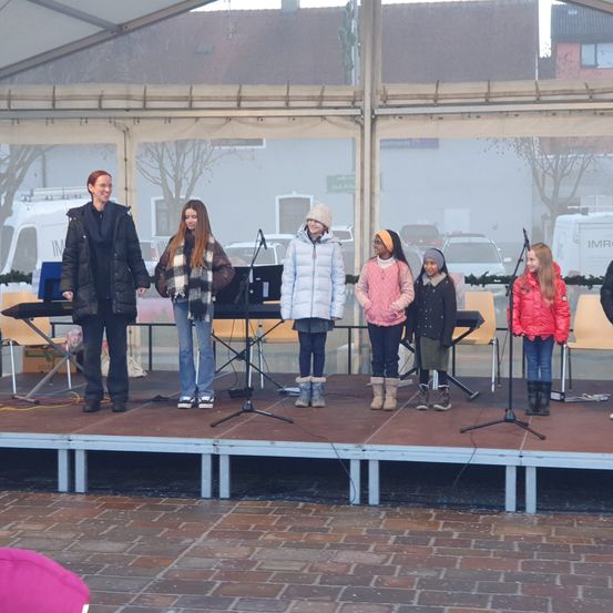 Eine Gruppe von Kindern steht auf einer Bühne unter einem Zelt mit einer erwachsenen Frau. Sie haben Musikinstrumente und scheinen sich auf eine Aufführung vorzubereiten.