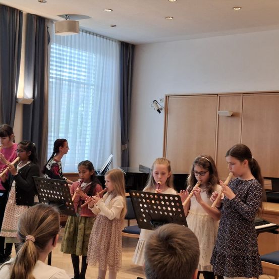 Eine Gruppe junger Mädchen spielt Flöten in einem Raum mit einem Klavier. Eine Frau in einem rosa Shirt schaut zu.