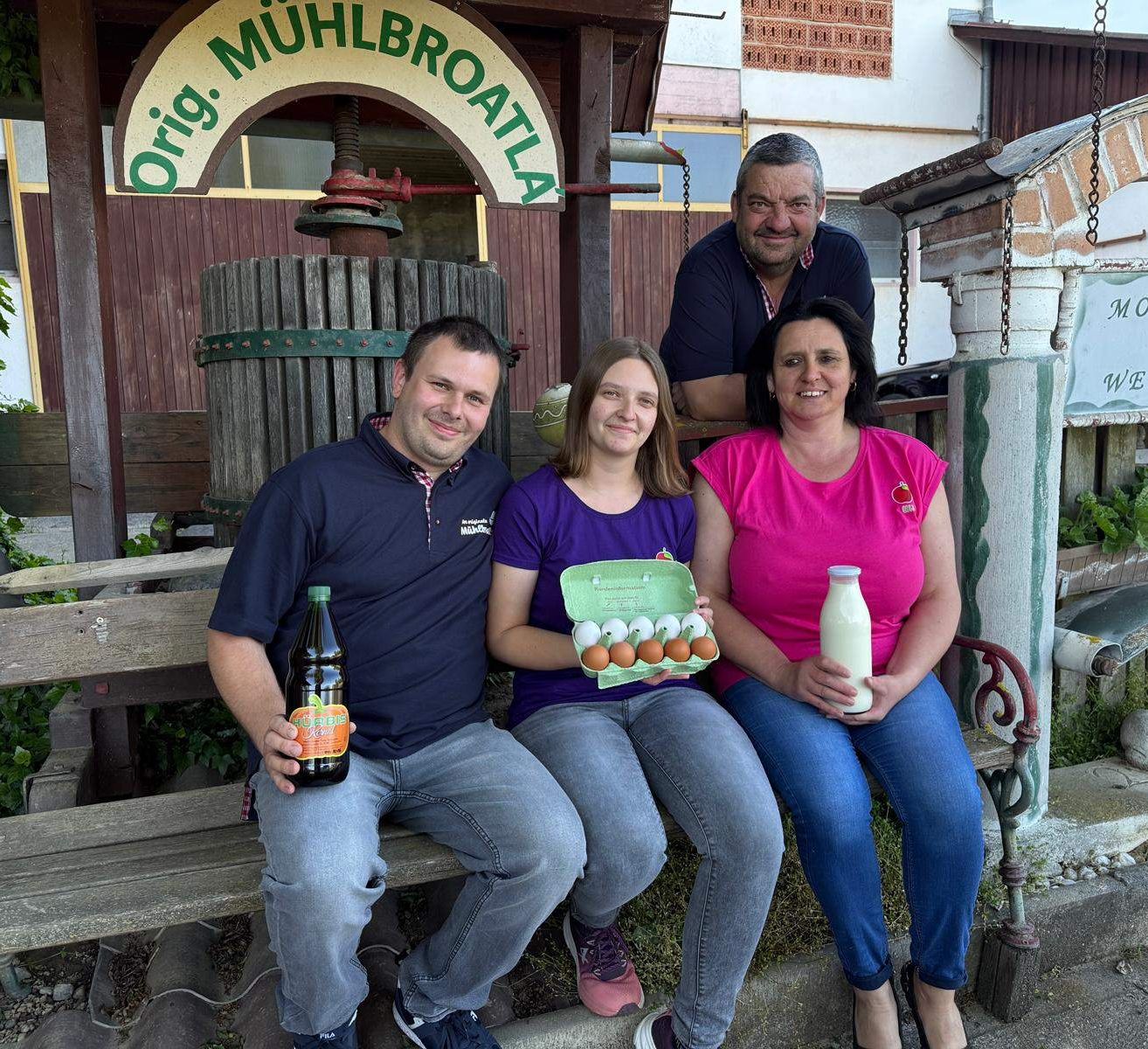 Vier Personen lächeln und posieren für ein Foto vor einem Holzgebäude mit einem Schild, das 'Orig Mühlbrota' lautet. Ein Mann auf der linken Seite hält eine Flasche Bier. Eine Frau in der Mitte hält ein Karton mit Eiern, und eine Frau auf der rechten Seite hält eine Flasche Milch. Hinter ihnen befindet sich eine große Holzpresse.