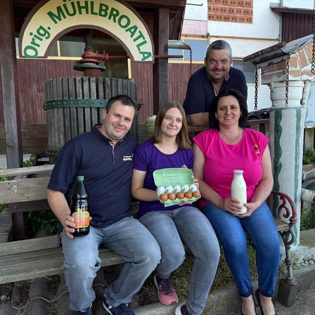 Vier Personen lächeln und posieren für ein Foto vor einem Holzgebäude mit einem Schild, das 'Orig Mühlbrota' lautet. Ein Mann auf der linken Seite hält eine Flasche Bier. Eine Frau in der Mitte hält ein Karton mit Eiern, und eine Frau auf der rechten Seite hält eine Flasche Milch. Hinter ihnen befindet sich eine große Holzpresse.