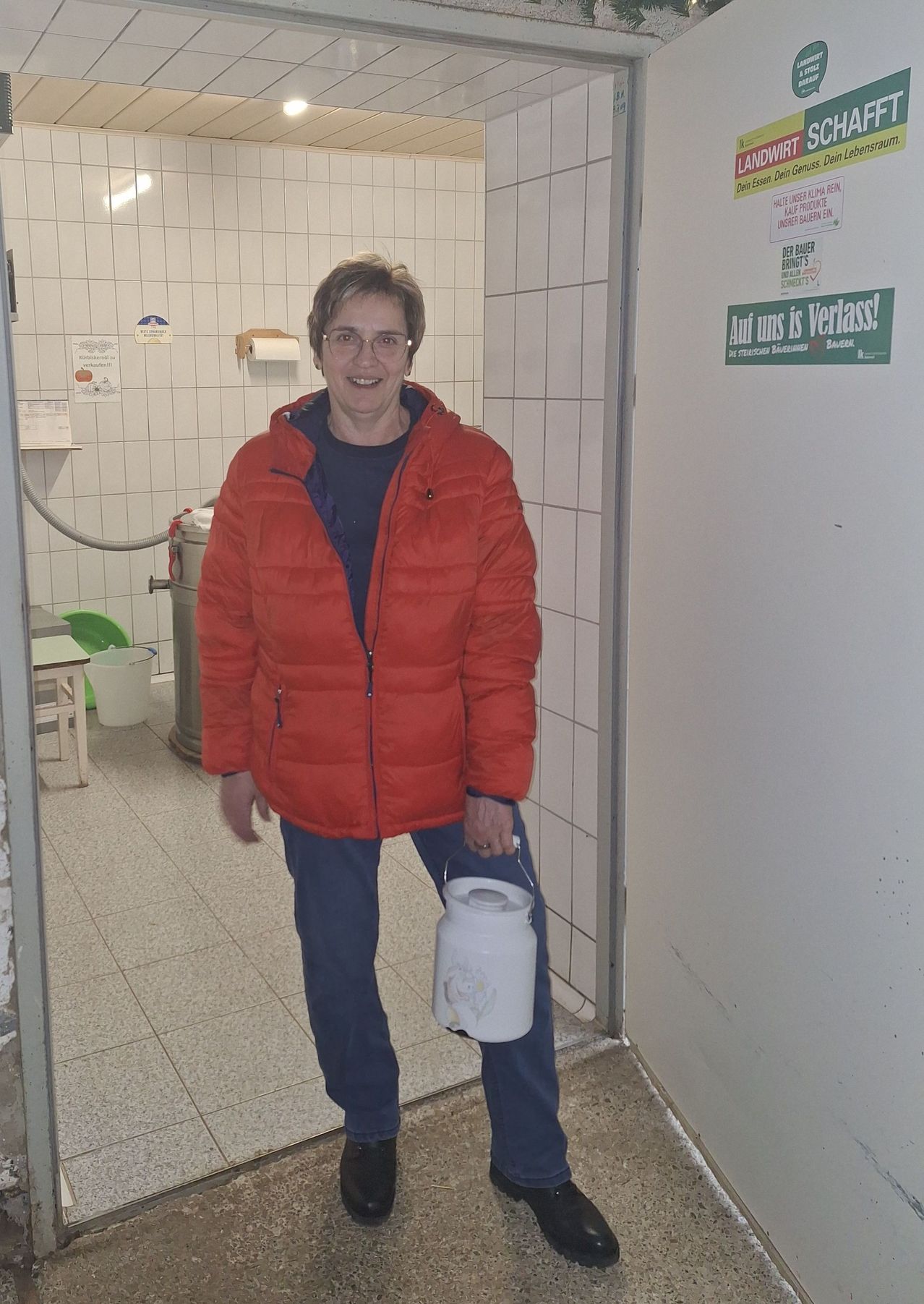 Eine Frau mit Brille und einer roten Jacke steht lächelnd in einem Badezimmer und hält einen weißen Eimer. Das Badezimmer hat weiße Fliesen und ein grünes Schild an der Wand.