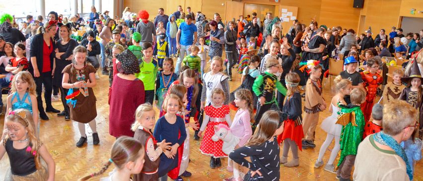 Eine große Gruppe von Kindern in Kostümen versammelt sich in einem Raum, einige stehen, andere gehen herum. Der Raum ist mit Konfetti auf dem Boden dekoriert.
